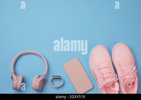 un paio di sneaker rosa, cuffie wireless, uno smartphone e un orologio intelligente su sfondo blu. Oggetti e gadget per lo sport, la corsa, la posa piatta Foto Stock