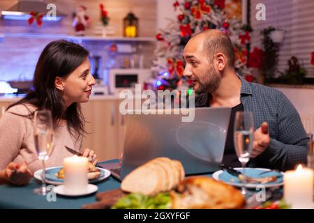 Coppia gioiosa che guarda film di intrattenimento sul computer portatile mentre si siede al tavolo da pranzo in cucina decorata con Natale. Famiglia felice festeggiamo le vacanze di natale godendo insieme la stagione invernale Foto Stock