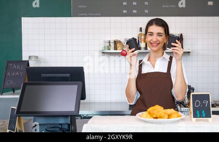 Il personale di una caffetteria caucasica giovane si vestiva di grembiule marrone e teneva una bevanda a base di caffè in una tazza di plastica con entrambe le mani. Atmosfera mattutina in una caffetteria. Foto Stock