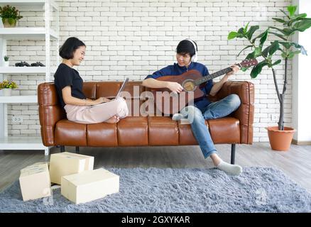 I giovani amanti trascorrono il tempo insieme in vacanza nel soggiorno. Uomo asiatico con le cuffie che suonano una chitarra acustica mentre la ragazza controlla il pacco Foto Stock