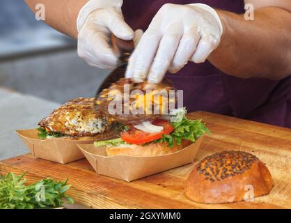 Uno chef prepara un hamburger per un cliente. Un uomo che indossa guanti protettivi bianchi raccoglie gli hamburger dalla griglia e li aggiunge alla pistola per hamburger. MO Foto Stock