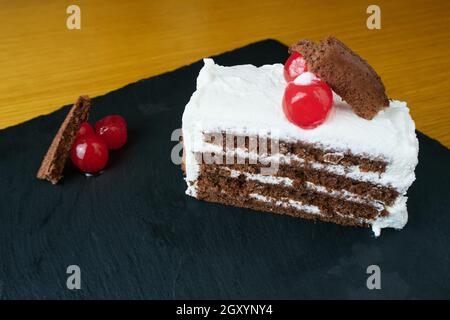 Fetta di foresta nera decorata con ciliegie e piccoli pezzi di torta su una tavola di ardesia nera Foto Stock