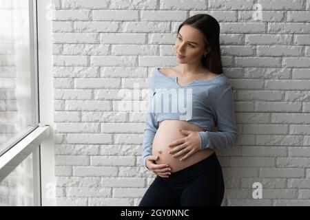 Ritratto di una giovane donna incinta. Si alza contro un muro di mattoni e guarda fuori la finestra. Foto Stock