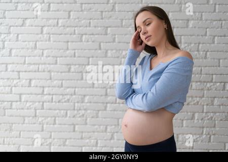 Una donna si erge vicino ad un muro di mattoni bianchi. Tiene la testa con la mano. Concetto di gravidanza esperienza Foto Stock