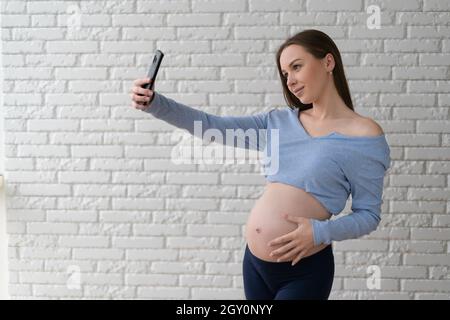 Una donna si alza vicino fa un selfie sul telefono. Ritratto di una giovane incinta Foto Stock