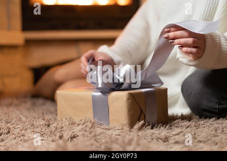 Una giovane donna siede sul pavimento accanto al camino e scollega un arco su una scatola con un regalo. Il concetto di vacanza e sorprese. Foto Stock