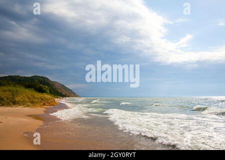 Paesaggio di una spiaggia sabbiosa con onde bianche e schiumose che lambiscono in lontananza le erbe e la montagna Foto Stock