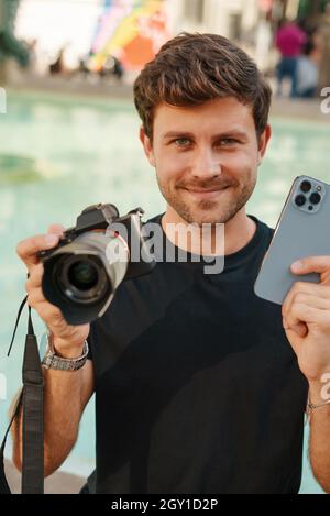 Felice giovane fotografo maschio sorridente e mostrando contemporaneo telefono cellulare e fotocamera digitale con obiettivo ottico, mentre si siede vicino alla fontana nel parco della città Foto Stock