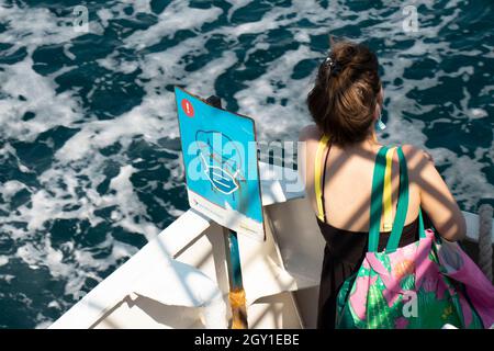 Vodice, Croazia - 25 agosto 2021: Indossare il cartello con la maschera COVID-19 e una giovane donna passeggero che guarda l'acqua di mare da una nave in movimento, angolo alto v Foto Stock