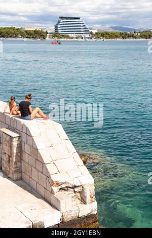 Vodice, Croazia - 25 agosto 2021: Persone che siedono su un molo di pietra vicino al mare e moderno edificio alberghiero Olympia Sky in lontananza, alto angolo di vista Foto Stock