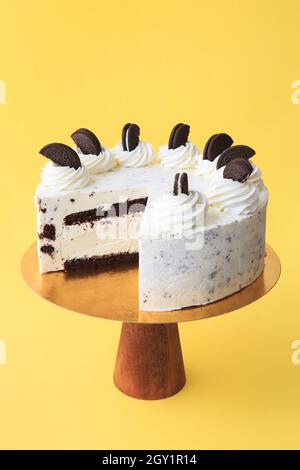 Fetta torta di compleanno sul supporto per torta di legno. Bella torta bianca con panna montata decorata con biscotti al cioccolato nero. Sfondo giallo. Copia Foto Stock