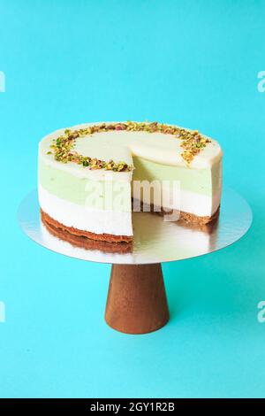 Fetta torta di compleanno sul supporto per torta di legno. Bella torta al pistacchio con panna montata. Sfondo turchese. Spazio di copia. Fotografia alimentare per rec Foto Stock