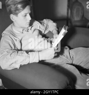 Ein junger Mann sitzt in einem Sessel und liest ein Buch, Deutschland 1940. Un giovane uomo seduto in una sedia la lettura di un libro, Germania 1940. Foto Stock