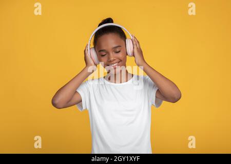 Soddisfatto carino adolescente afro american ragazza in bianco t-shirt in cuffie ascolto musica Foto Stock