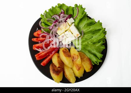 Piatto nero pieno di patate al forno, formaggio fresco, pomodori a fette, cipolla rossa a fette, foglia di lattuga e olio d'oliva. Cibo mediterraneo isolato su wh Foto Stock