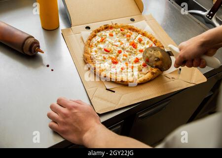 Una pizzaiolo taglia una pizza con un coltello rotondo prima di avvolgersi per la consegna. Le mani degli uomini di fronte alla pizza. Foto Stock