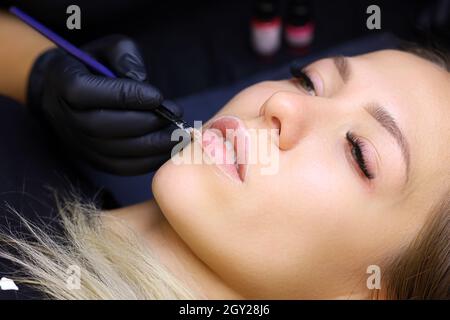 il padrone tiene un pennello nelle sue mani e pulisce il contorno delle labbra del modello prima della procedura di trucco permanente del labbro Foto Stock