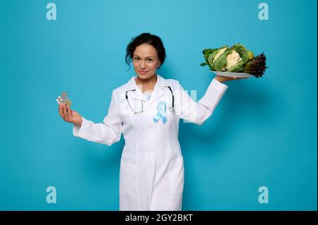 Bel medico sicuro in abito medico bianco con nastro blu, simbolo della Giornata Mondiale della consapevolezza del diabete, tiene un piatto con sano vegano mangiare e. Foto Stock