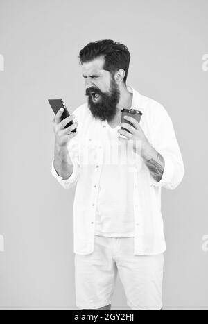 arrabbiato uomo maturo bearded gridare emozionalmente sul telefono mentre beve il caffè del mattino, esprimendo la rabbia Foto Stock