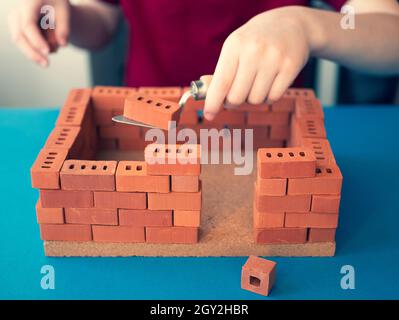 Il bambino sta generando una costruzione con i mattoni del giocattolo e la cazzuola della mano. Concetto della professione di ingegnere strutturale, costruttore. Fare una casa giocattolo Foto Stock