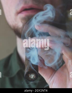 Un uomo accende un sigaro cubano con un accendisigari. Un giovane uomo in una maglietta verde sta fumando vicino alla finestra. Primo piano Foto Stock