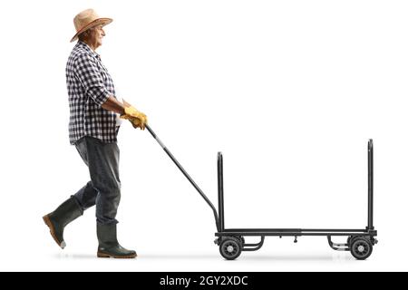 Colpo di profilo a tutta lunghezza di un agricoltore maturo che cammina e spinge un camion a mano vuoto isolato su sfondo bianco Foto Stock