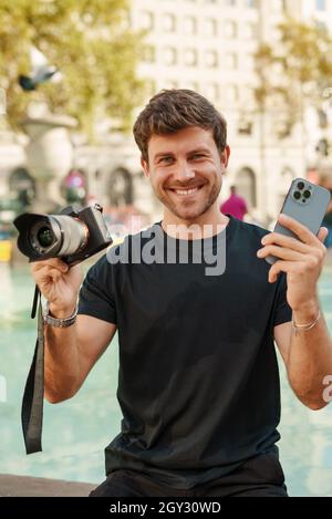 Felice giovane fotografo maschile in abiti casual sorridendo, mentre dimostra la moderna fotocamera DSLR e smartphone seduto vicino alla fontana nella giornata di sole nel parco della città Foto Stock