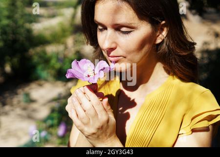 Attraente giovane spagnola donna che odora fresco viola ibisco fiore in un parco Foto Stock