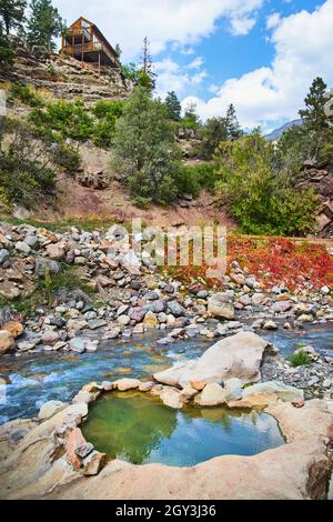 Piccola sorgente termale naturale vicino al fiume e scogliere con capanna di legno in cima Foto Stock