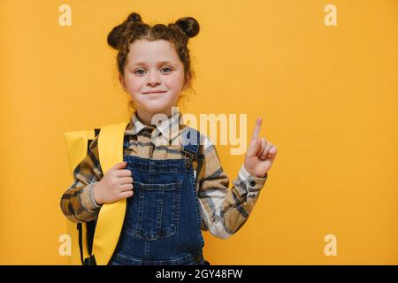 Felice carino bambino scuola elementare primaria ragazza con zaino puntando dito da parte a copia spazio pubblicità prodotti o servizi per gli allievi Foto Stock