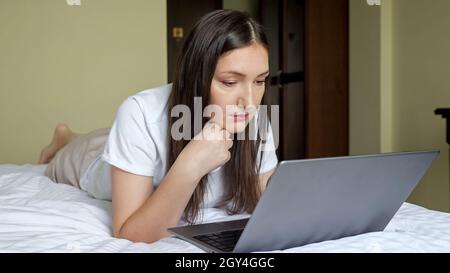 donna bruna che lavora su un computer portatile sdraiato sullo stomaco a letto Foto Stock