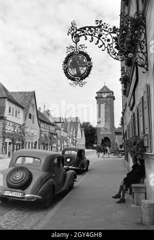 Zwei Schuljungen, die auf einer Bank vor zwei Autos sitzen, schauen einem älteren Verkehsmittel, der Pferdekutsche hinterher, Rothenburg ob der Tauber, Deutschland 1930er Jahre. Due scolari seduta su una panchina di fronte due vetture, sta guardando un molto più vecchio veicolo passando da una carrozza trainata da cavalli, Rothenburg ob der Tauber, Germania 1930s. Foto Stock