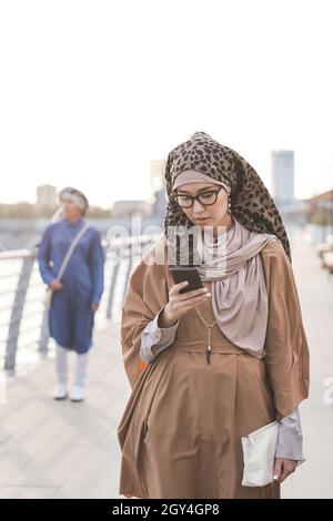 Giovane musulmana che comunica un messaggio sul suo cellulare mentre cammina in città Foto Stock