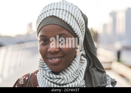 Primo piano della giovane donna africana in abiti musulmani sorridenti alla macchina fotografica mentre si trova all'aperto Foto Stock
