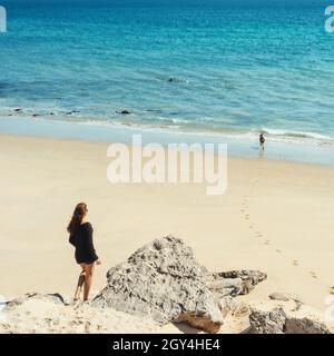 Idilliaco alto angolo vista mare con una donna solitaria a piedi sulla spiaggia soleggiata con il suo cane giocoso godendo una giornata estiva sulla riva del mare Foto Stock