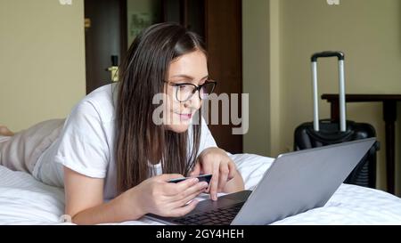 donna in occhiali con una carta funziona su un computer portatile, sdraiato sul letto sullo stomaco contro lo sfondo di una valigia Foto Stock