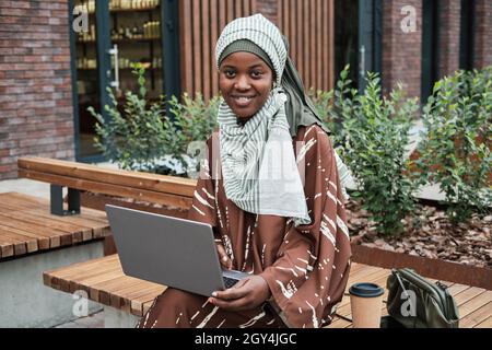 Ritratto della donna d'affari musulmana africana seduta sul banco con il portatile all'aperto e sorridente alla macchina fotografica Foto Stock