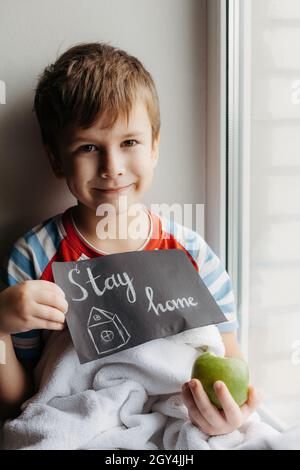 Ragazzino seduto a casa in quarantena. Il bambino tiene in mano un segno con l'iscrizione Stay Home Foto Stock