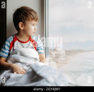 Ragazzino triste che sii a casa in quarantena. Un bambino malato si siede vicino alla finestra e guarda al mare Foto Stock