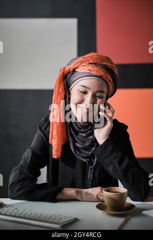 Donna d'affari di successo in abbigliamento musulmano seduto sul suo posto di lavoro in ufficio prendendo il telefono cellulare e sorridendo Foto Stock