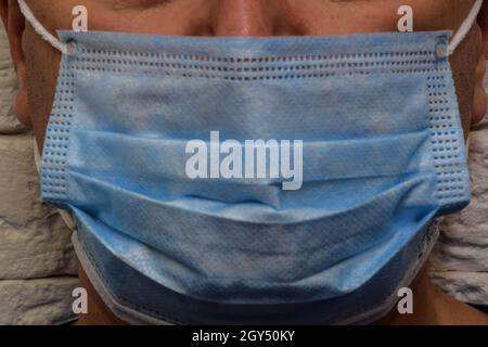 Maschera medica blu su un maschio faccia close-up contro uno sfondo muro di mattoni. Viene rimossa solo la metà inferiore della faccia Foto Stock