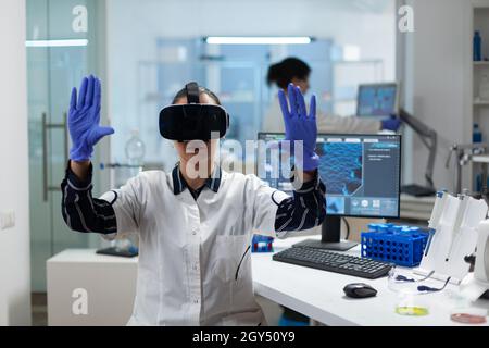 Ricercatore biologo che indossa cuffie per realtà virtuale esaminando esperimento di biochimica che lavora in laboratorio ospedaliero di microbiologia utilizzando biotecnologie. Scienziato specializzato che analizza lo sviluppo del virus Foto Stock