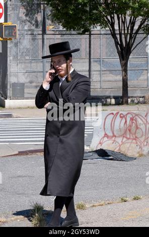 Un giovane ebreo hasidico con un lungo peyus riccio parla sul suo mobile. Su Division Avenue a Williamsburg, Brooklyn, New York. Foto Stock