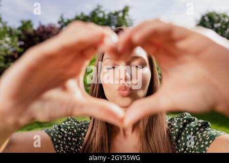 Foto di flirty bella felice ragazza allegra fare dita cuore forma inviare aria bacio all'aperto fuori parco Foto Stock