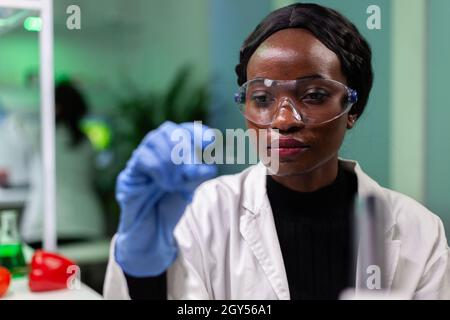 Primo piano di donna biochimica afroamericana che tiene campione di foglie verdi lavorando all'esperimento medico in laboratorio di ospedale di microbiologia. Medico botanico che analizza piante geneticamente modificate Foto Stock