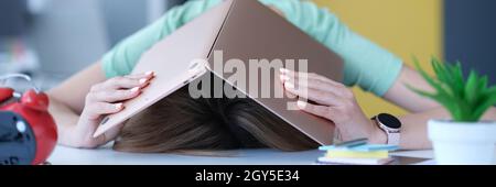 Stanca nello stress, la donna nasconde la testa con un computer portatile sul posto di lavoro. Foto Stock