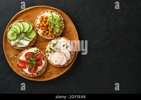 Quattro croccanti pane di grano saraceno con formaggio spalmabile, rafano, pomodoro, ceci, cetrioli e microsverde per una sana colazione su carta pergamena su cartone nero Foto Stock