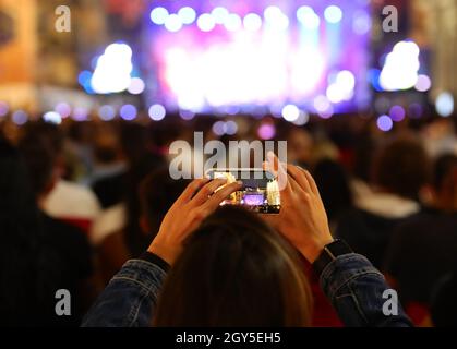 ragazza che scatta un video con il tuo smartphone durante un concerto dal vivo con molto pubblico e il palco con molte luci Foto Stock