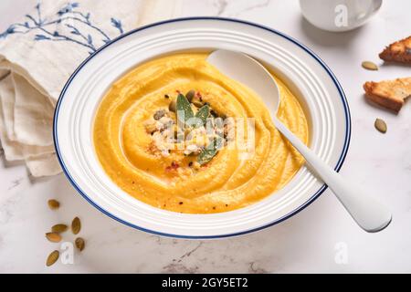 Zuppa tradizionale di zucca con consistenza cremosa e setosa con foglie di salvia e pepe rosso in piatto bianco o ciotola. Sfondo grigio chiaro. Spazio di copia. Mock up. Foto Stock