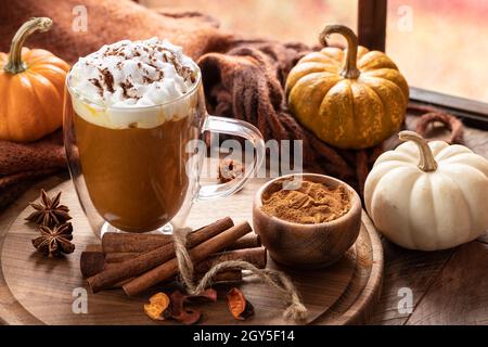 Latte di zucca speziata con bastoncini di cannella su un piatto di legno e zucche da una finestra Foto Stock